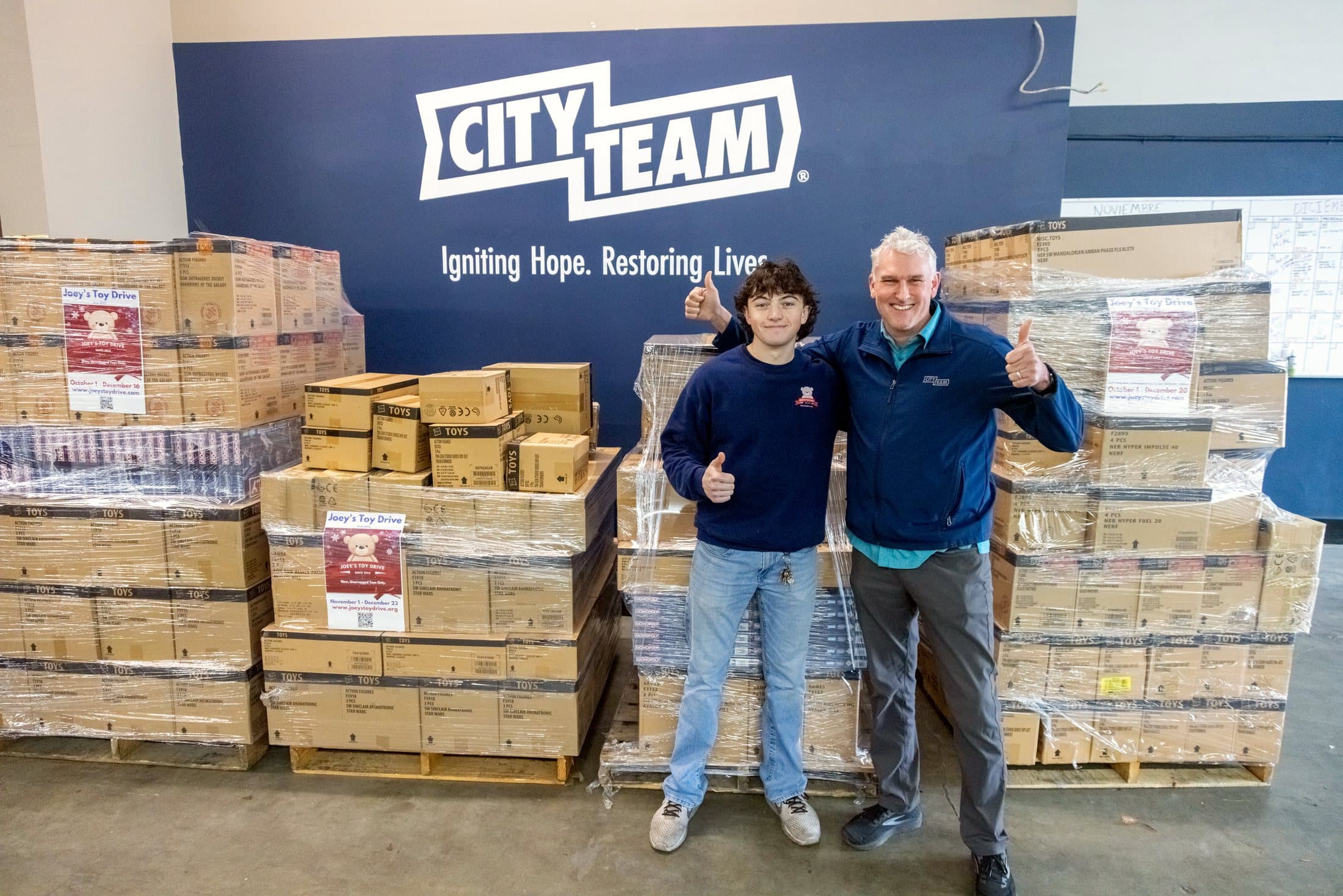 Pallets of donated toys stacked under a CityTeam sign, with two people standing in front of them