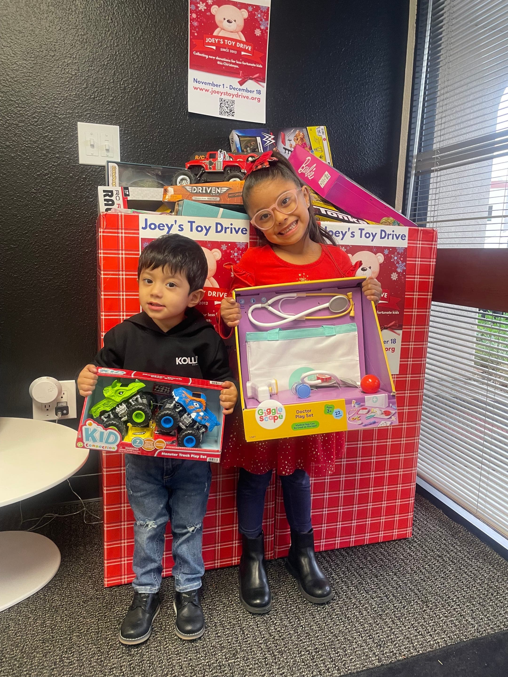 Two children holding the new toys they just received next to a Joey's Toy Drive donation box