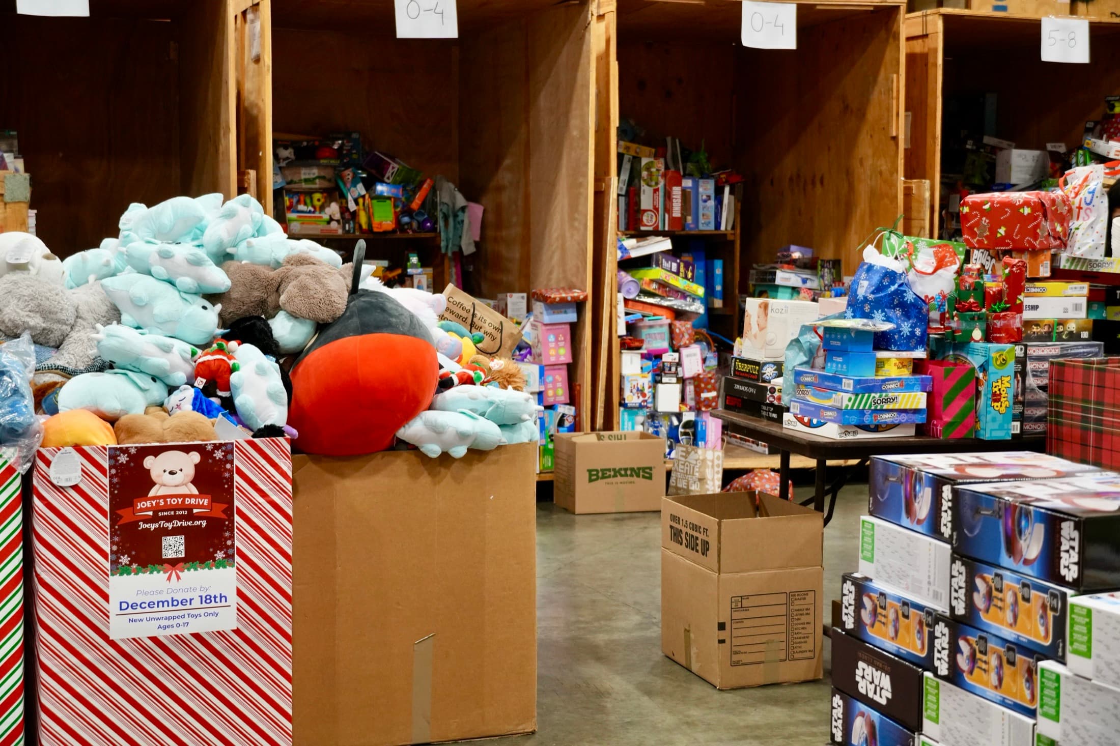 A Joey's Toy Drive collection box beside bins of stuffed animals and stacked toys inside the warehouse