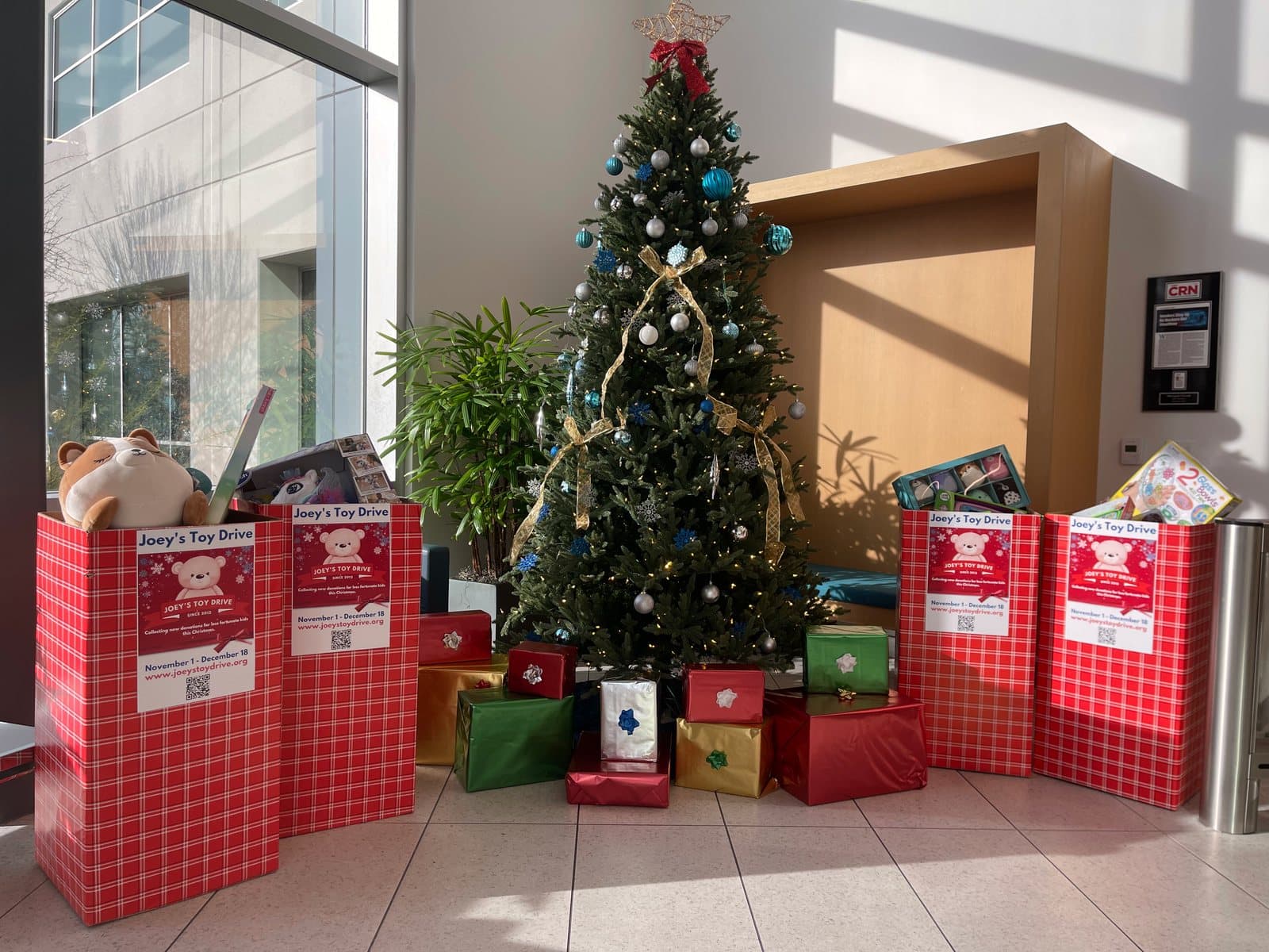 Four Joey's Toy Drive collection boxes standing around a decorated Christmas tree in an office lobby